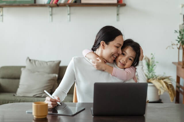 A woman working at a laptop whilst being hugged by a child